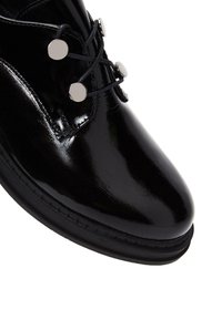 Zwarte patentleren schoen met een ronde neus, voorzien van zilveren ronde accenten en vetersluiting aan de voorkant. Gladde, glanzende afwerking.
