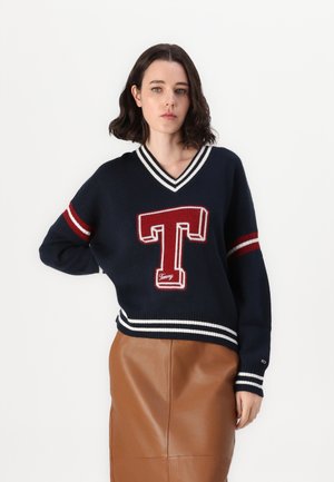 Maglione a maglia navy con scollo a V, caratterizzato da polsini e colletto a righe rosse e bianche, e un grande design ricamato a forma di "T" sulla parte anteriore.