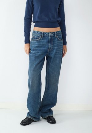 Person iført løstsiddende blå jeans, en kort marineblå sweater og sorte sko står op ad en almindelig hvid væg.