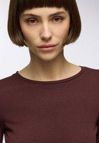 Burgundowa bluzka o gładkiej teksturze i okrągłym dekolcie, z dopasowanym krojem i krótkimi rękawami. Tło jest neutralne.