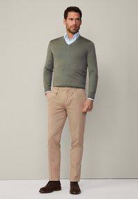 Un pull vert clair en V sur une chemise bleue à col, associé à un pantalon beige et des bottes marron, se tenant contre un fond neutre.