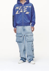Sudadera con cremallera azul que lleva el texto "NUNCA ES TARDE", combinada con unos pantalones cargo de corte holgado y lavado claro con múltiples bolsillos y zapatillas blancas.
