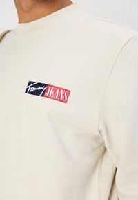 Beige genser med ribbestrikket hals, med et fremtredende rektangulært logo i rødt og blått som sier "Tommy JEANS" på venstre bryst.