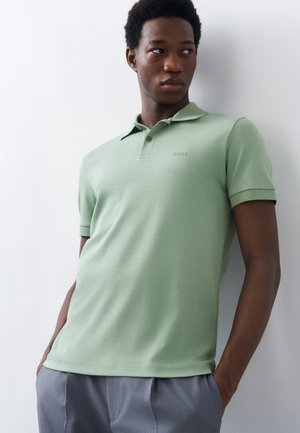 Ung mann i lysegrønn kortermet poloskjorte og grå bukser, stående med hendene i lommene, ser sidelengs mot en hvit vegg.