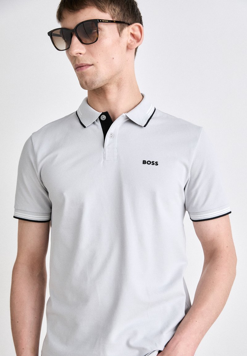 Polo shirt gris clair avec col et accents de manches noirs, arborant un petit logo noir. Porté avec des lunettes de soleil noires. Texture lisse.
