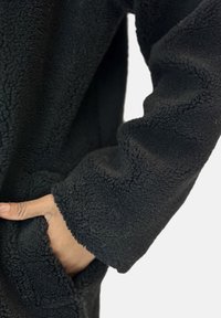 Schwarze Fleecejacke mit strukturierter Oberfläche, die eine Hand in einer Seitentasche zeigt. Die Bündchen sind übergroß und leicht umgeschlagen.
