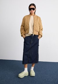 Veste bomber matelassée marron, t-shirt beige, jupe en denim foncé et sabots vert clair portés avec des chaussettes blanches ; chaussures plates, style décontracté.