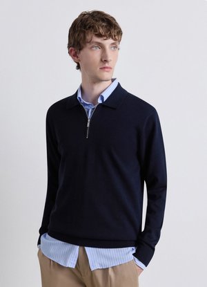Junger Mann mit welligem braunem Haar, der einen marineblauen Reißverschluss-Pullover über einem hellblau gestreiften Hemd und beigen Hosen trägt und zur Seite schaut.