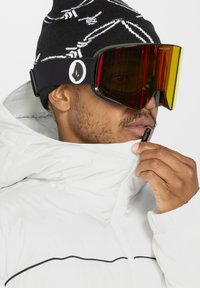 Skibril met grote, gebogen, rood getinte lenzen. Zwarte elastische band met wit logo. Model draagt een witte, met rits sluitende jas en een zwarte muts.