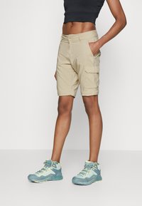 Beige Cargo-Shorts mit Seitenpockets, aus leichtem Stoff. Kombiniert mit hellblauen High-Top-Sneakers mit gelben Akzenten.