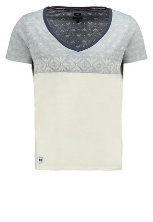 Zwarte bomberjack met rits en grijze mouwen, gedragen over een wit shirt met een geometrisch patroon. Gecombineerd met donkerblauwe jeans en witte instapschoenen.