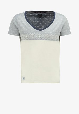 Veste bomber noire zippée avec des manches grises, portée sur une chemise blanche à motif géométrique. Associée à un jean bleu foncé et des chaussures blanches à enfiler.