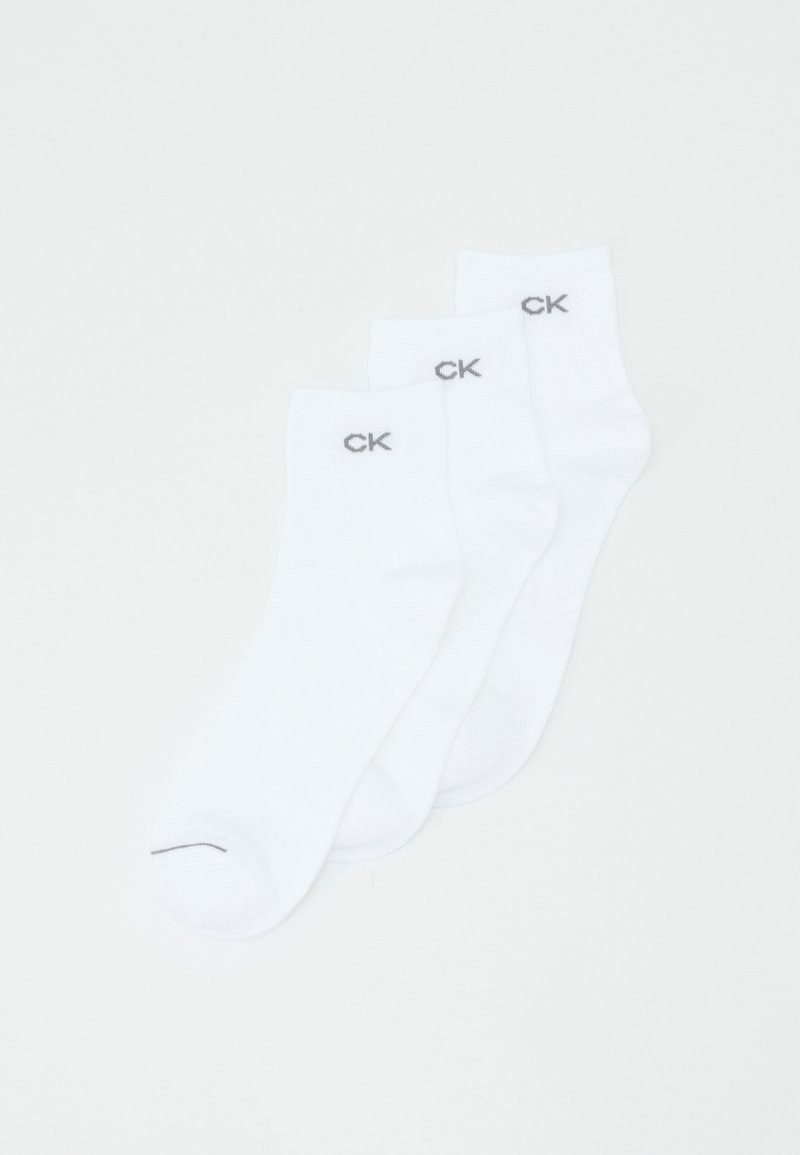 Tre par hvide bomuldssokker udstillet, med en ribbet kant og grå "CK" logo nær toppen. Glat tekstur og klassisk ankel længde.