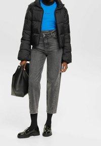 Sort pufferjakke, blå top med tre prikker, højhalsede grå jeans, sorte loafers og en sort totebag. Glatte strukturer hele vejen igennem.