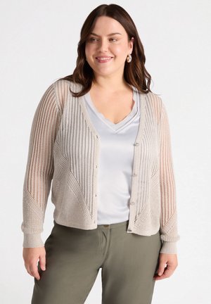 Cardigan en tricot beige clair avec un tissage ouvert et un design structuré ; comprend une fermeture à boutons et des manches longues, associé à un haut gris et un pantalon olive.