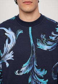 Granatowy sweatshirt z wyraźnym kwiatowym wzorem w jasnoniebieskim kolorze, z prążkowanym dekoltem i gładką fakturą. Zbliżenie na okolicę klatki piersiowej.