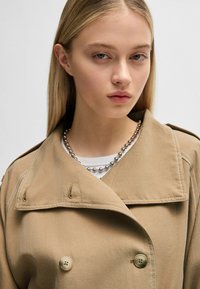 Beige trench coat with double-breasted design, large collar, and gold hardware accents. Features silver bead necklace and white shirt underneath.