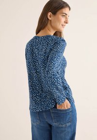 Blau gemusterter Langarmoberteil mit kleinen weißen und hellblauen Formen, getragen mit hellblauen Jeans aus Denim mit Hintertaschen.