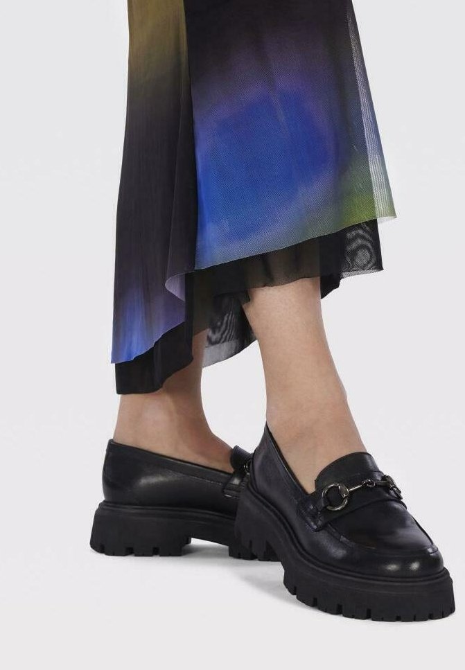 Mocassins en cuir noir avec une semelle épaisse et un accent chaîne, associés à une jupe fluide multicolore dégradée avec une superposition transparente.