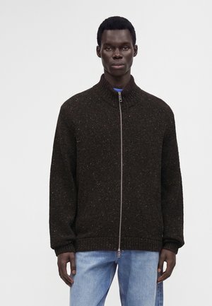 Homme portant un pull en maille marron foncé à fermeture éclair et un jean bleu clair, debout devant un fond blanc uni.