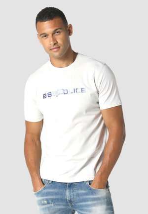 White short-sleeved t-shirt with a blue metallic "883 Police" logo across the chest; modeled with blue distressed jeans.