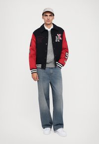 Jachetă varsity neagră și roșie cu dungi albe, pulover gri cu tricotaj în relief, blugi albaștri cu croială largă și adidași albi. Șapcă gri cu logo.