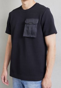 Svart t-shirt i bomull, med en framficka med klaff, logotypdetalj och korta ärmar, matchad med ljusa denimjeans.