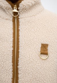 Veste en polaire couleur crème avec bordure marron, fermeture éclair dorée et une petite boucle en tissu marron avec un anneau doré sur la poitrine.