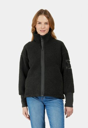 Kvinde iført en sort fleecejakke med lynlås og høj krave samt blå jeans, står mod en ensfarvet lys baggrund og vender fremad.