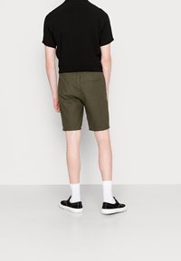 Shorts vert olive avec une poche arrière, portés avec une chemise noire à manches courtes et des chaussures noires, assortis à des chaussettes blanches montantes.