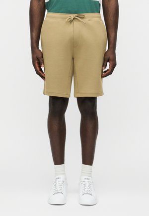 Mężczyzna ubrany w beżowe szorty z troczkami, zielono-biały pasiasty shirt, białe skarpety i białe sneakersy Polo stoi na jednolitym tle.