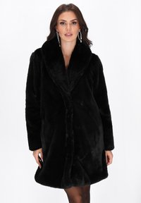 Mujer vestida con un abrigo largo de piel sintética negra con un gran cuello, combinado con grandes pendientes de aro de cristal, contra un fondo blanco liso.