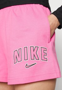 Růžové šortky Nike vyrobené z měkké tkaniny s elastickým pasem. Obsahují výrazný černobílý potisk loga "NIKE" na lemu a postranní kapsy.