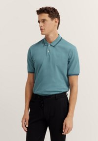 Polo di colore verde acqua con colletto, caratterizzato da doppie strisce navy. Maniche corte, logo discreto sul petto sinistro, abbinato a pantaloni scuri.