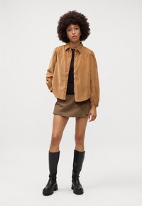 Camicia button-up in corduroy marrone sovrapposta a un top marrone, abbinata a una gonna mini a quadri e stivali neri fino al ginocchio.