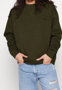 Sudadera verde oliva con puños y dobladillo acanalados, con el bordado "LE CLUB SPORTIF" en el pecho, combinada con vaqueros azul claro desgastados.