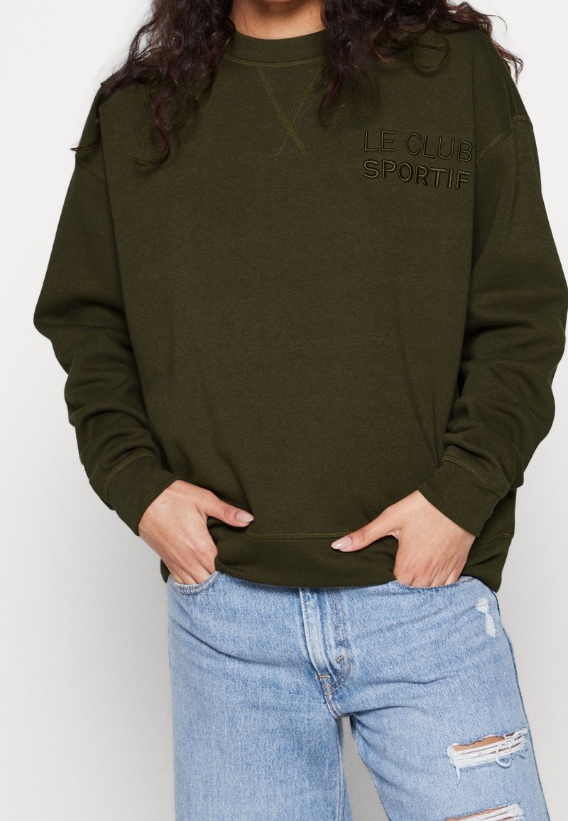 Sudadera verde oliva con puños y dobladillo acanalados, con el bordado "LE CLUB SPORTIF" en el pecho, combinada con vaqueros azul claro desgastados.