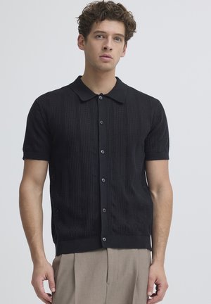 Junger Mann mit lockigem Haar, der ein schwarzes, kurzärmeliges Strickhemd mit Knöpfen und beige Hose trägt, steht vor einem schlichten Hintergrund.