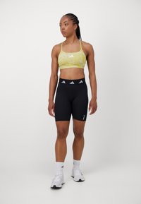 Sujetador deportivo amarillo con un patrón, con tirantes finos, combinado con pantalones cortos ajustados negros que presentan un logotipo blanco, y zapatillas deportivas blancas con cordones visibles.