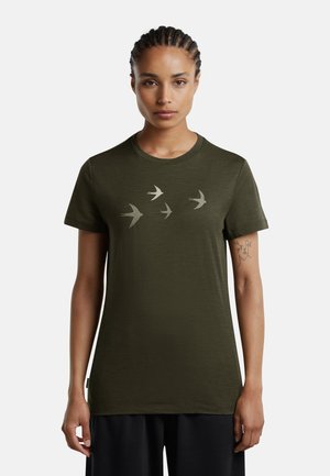 Kvinde med fletninger har en olivengrøn t-shirt med fire flyvende fuglesilhuetter over brystet og en tatovering på sin venstre arm.