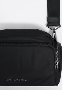 Čierna crossbody taška vyrobená z odolnej tkaniny, s textúrovaným povrchom, výrazným zipsom a logom embosovaným na prednej strane. Súčasťou je nastaviteľný popruh.