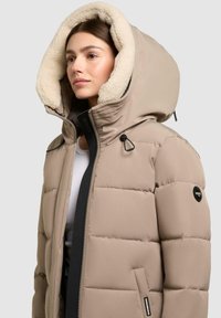 Giovane donna che indossa un cappotto invernale imbottito beige con un ampio cappuccio foderato in pile, chiuso con la cerniera, in piedi contro uno sfondo chiaro e semplice.