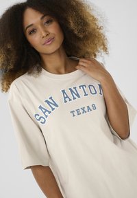 T-shirt oversize beige avec "SAN ANTONIO" en lettres bleues en gras et "TEXAS" en dessous. Manches courtes, tissu en coton doux, coupe décontractée.