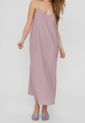 Kobieta w bezrękawnym maxi sukience w kolorze mauve i jasnoniebieskich sandałach z odkrytymi palcami, stojąca na jednolitym tle.