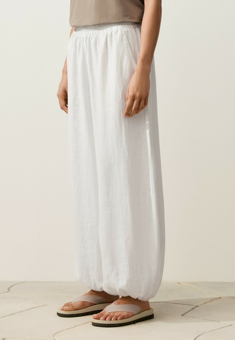 Personne portant un pantalon ample en lin blanc avec des chevilles élastiques et des sandales beiges, debout sur un sol carrelé.