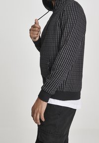 Hombre con sudadera negra y texto vertical blanco ajustando el cordón de la capucha, vistiendo pantalones negros y una camiseta blanca debajo.