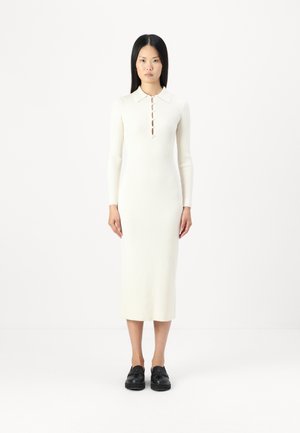 Prążkowana, kremowa sukienka z długim rękawem, z kołnierzykiem i zapięciem na cztery guziki. Modelka nosi czarne buty. Prosty, elegancki fason.