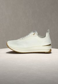 Λευκό χαμηλό αθλητικό παπούτσι με υφή πλεκτού, μπεζ σόλα, θηλιά στο πίσω μέρος και χρυσή λεπτομέρεια κοντά στη φτέρνα, τοποθετημένο σε ανοιχτόχρωμη επιφάνεια.