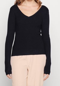 Maglione a V in blu navy scuro realizzato in morbido tessuto a maglia leggero; le maniche lunghe presentano fori per i pollici; abbinato a pantaloni beige chiaro.