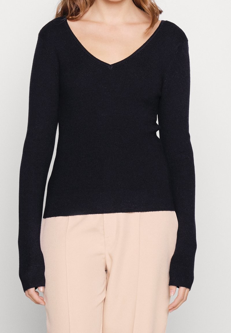 Maglione a V in blu navy scuro realizzato in morbido tessuto a maglia leggero; le maniche lunghe presentano fori per i pollici; abbinato a pantaloni beige chiaro.